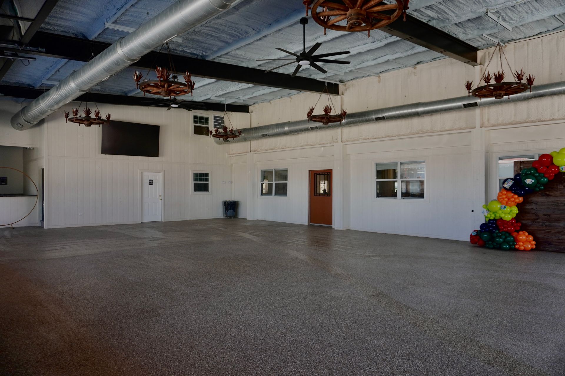 Empty indoor hall with white walls, gray floor, ceiling fans, and colorful stacked items in the corner