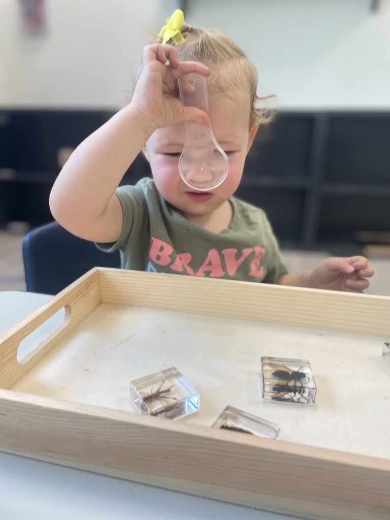 Child at a tray table, lowering a clear magnifying glass over small objects.
