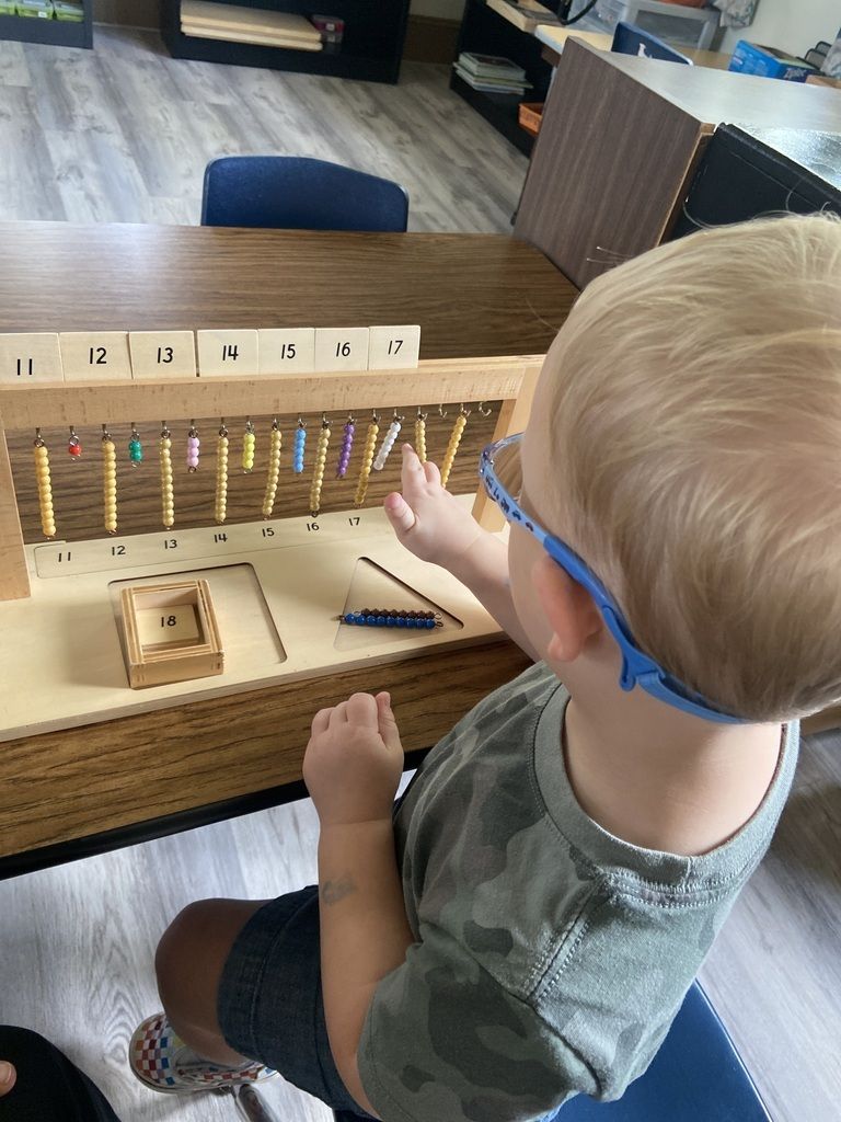 Child inspecting a wooden abacus at a table, reaching toward colorful beads in a classroom setting
