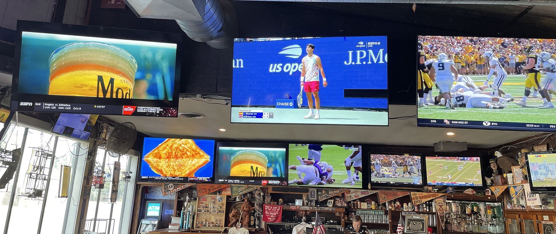 Televisions at a bar show beer, tennis, and football games.
