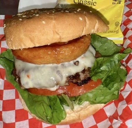 Exploded view of a burger with sesame seed bun, lettuce, tomato, cheese, patty, and red onion, with sauce splatters.