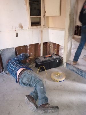 A person in jeans and a plaid shirt lies on the floor of a gutted kitchen near a toolbox and electrical cord.
