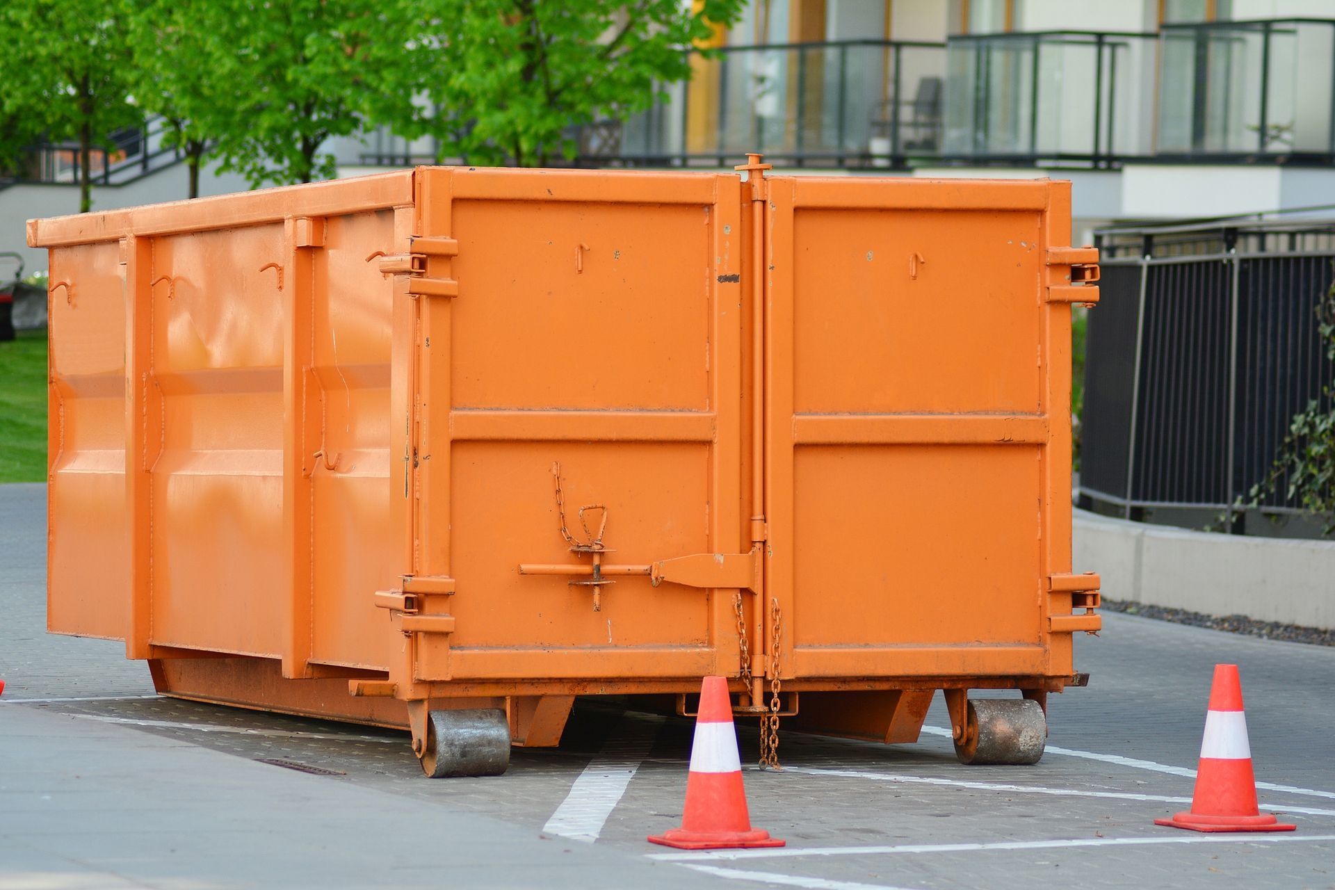 Orange dumpster on wheels in a parking area, with orange and white traffic cones placed in front of it.