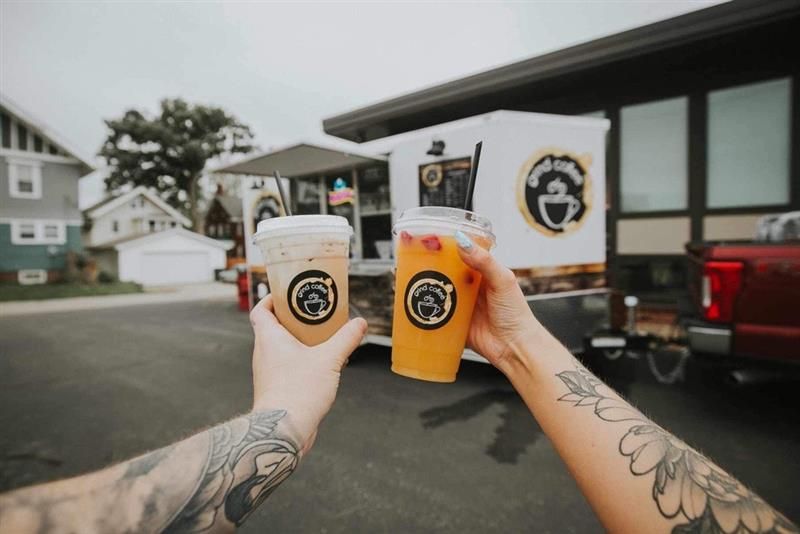 Hands holding iced drinks in front of a coffee truck, tattoos visible.