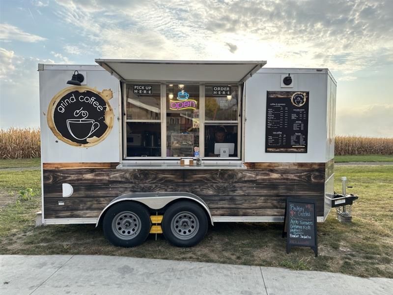 Coffee trailer with open window, menu board, and sign on a grassy area under a blue sky.
