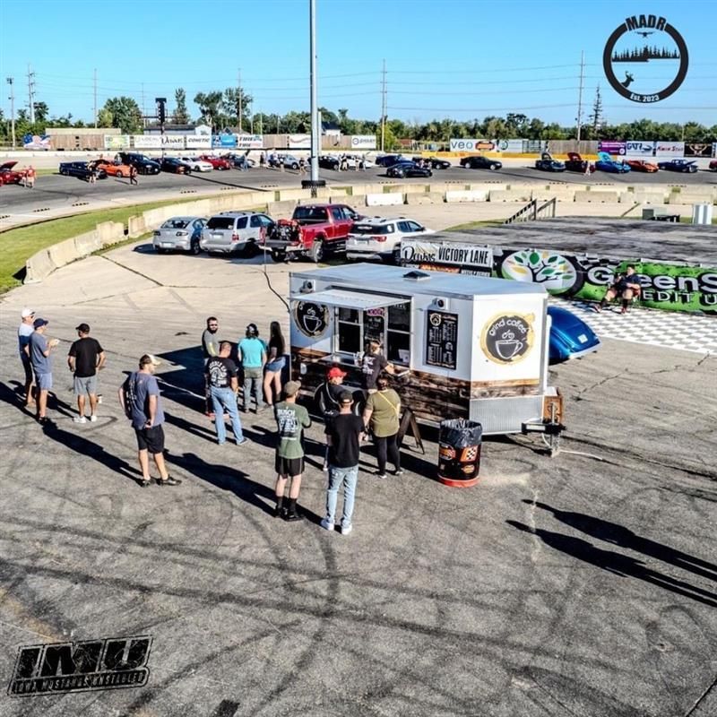A food truck with people gathers at a racetrack. Cars parked in the background.
