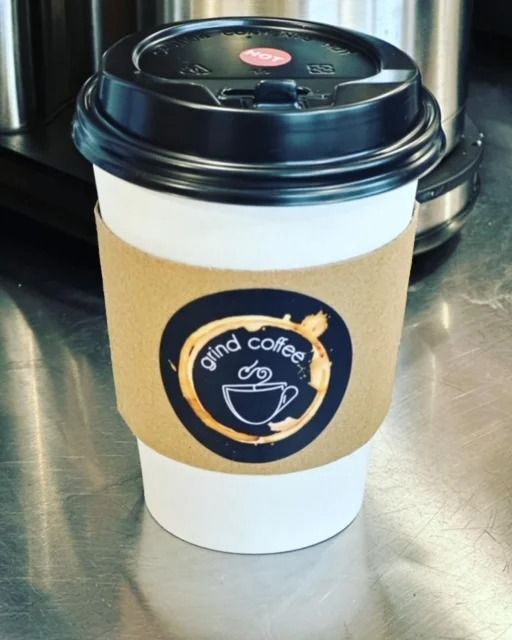 Coffee in a white cup with a brown sleeve, black lid, and "grind coffee" logo.