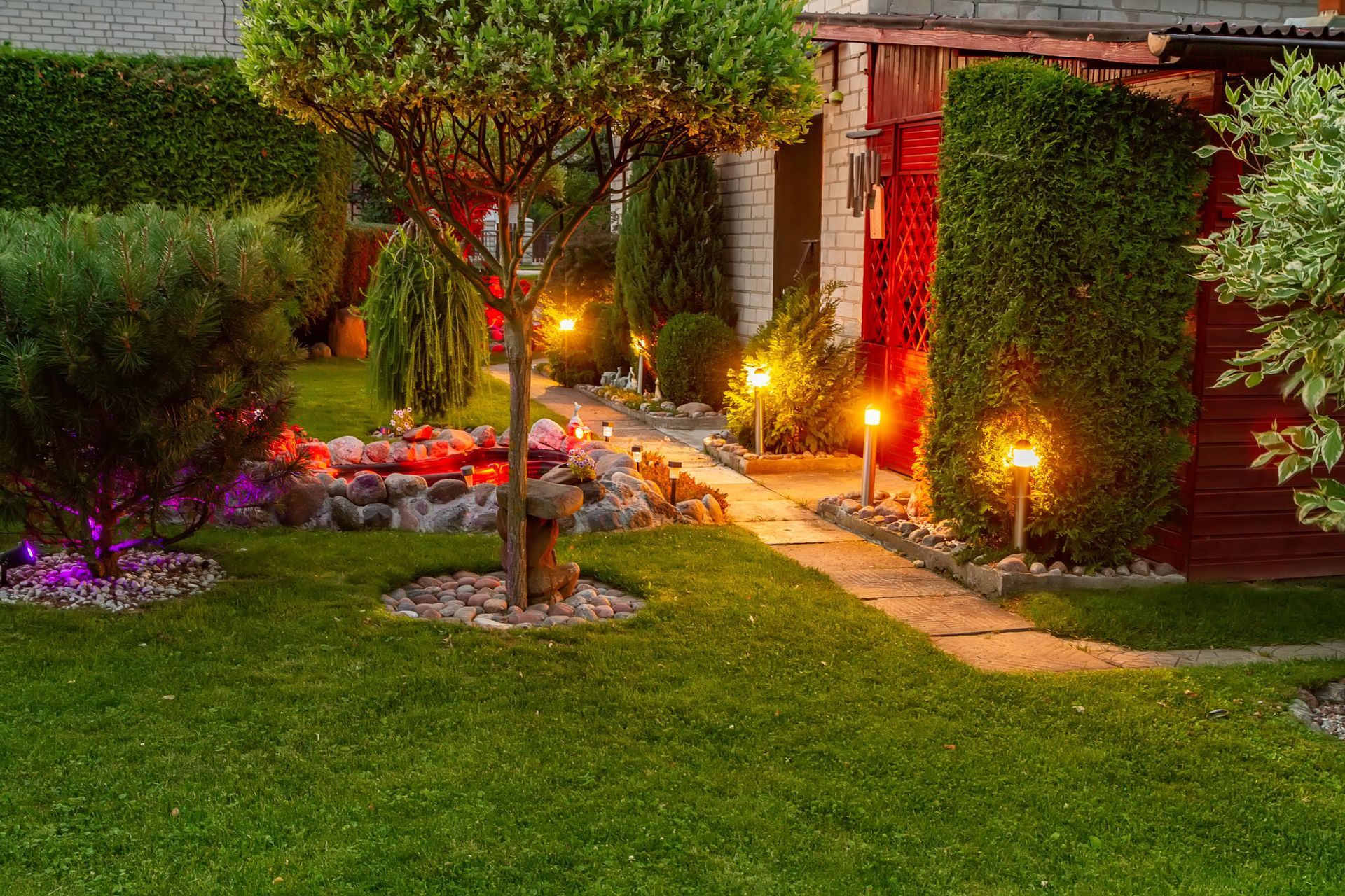 Lush backyard garden at night with pathway, lights, and trees.