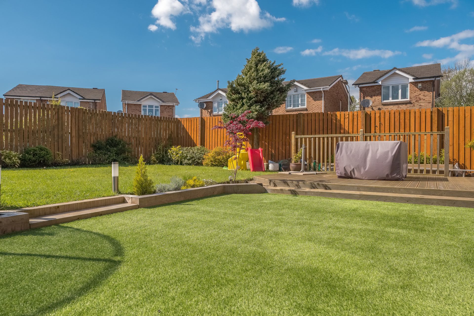 Backyard with green lawn, wooden fence, brick houses in the background, sunny day.