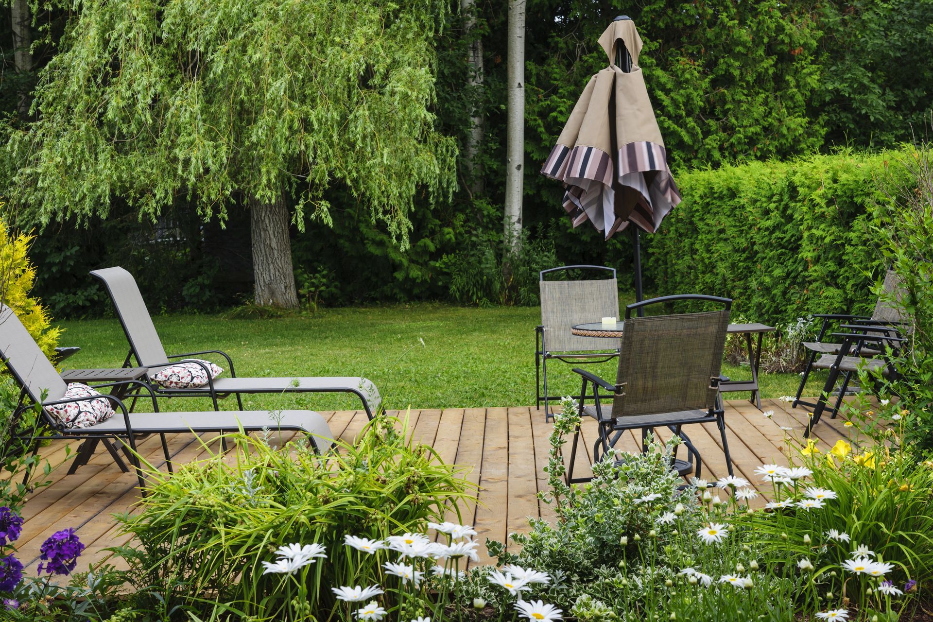 Wooden deck with lawn chairs, table, umbrella, and flowers. Lush green yard with trees.