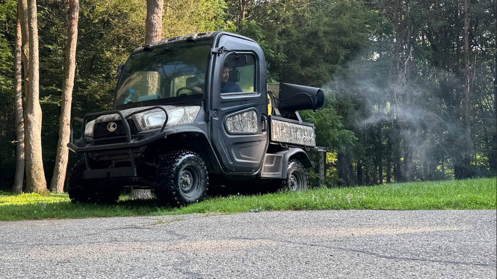 Dark utility vehicle spraying mist near a tree line on a gravel road.