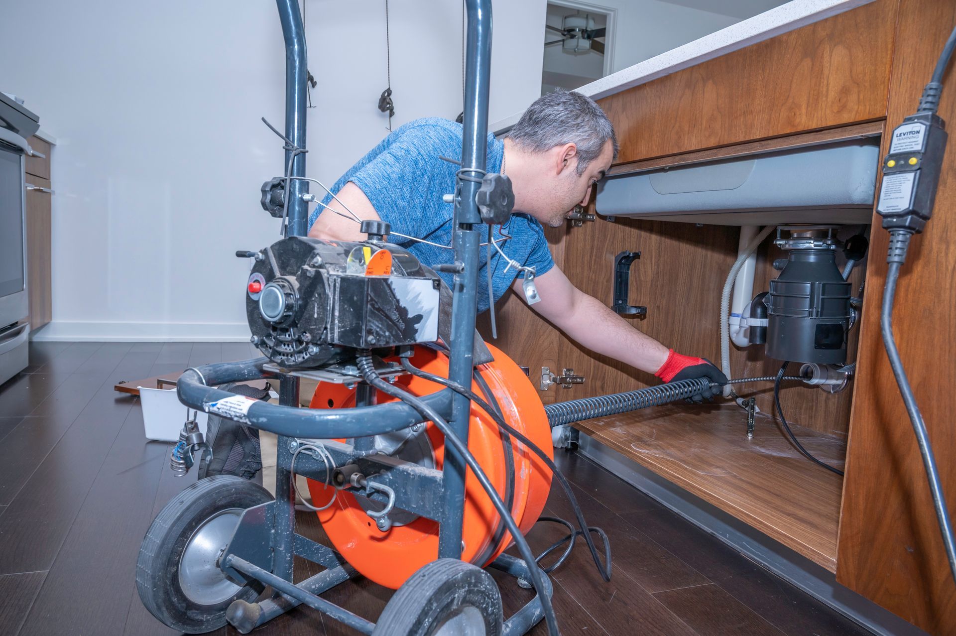 Plumber uses a drain snake under a kitchen sink. Grey machine, orange cable, brown cabinet, red gloves.