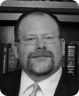 A middle-aged man with glasses and a goatee in a suit and tie, likely in a library or office setting.