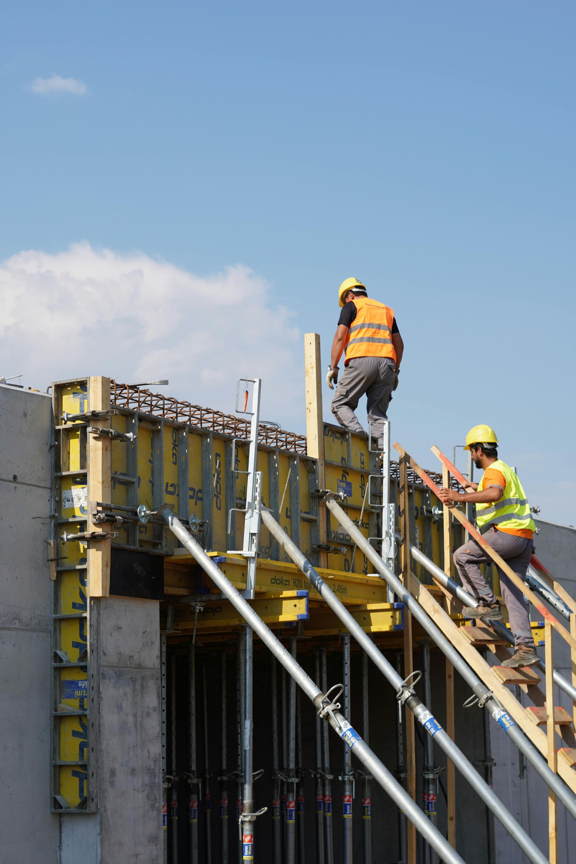 Construction workers on a partially built concrete structure, wearing safety vests and helmets. One is climbing, another is working on a stair.