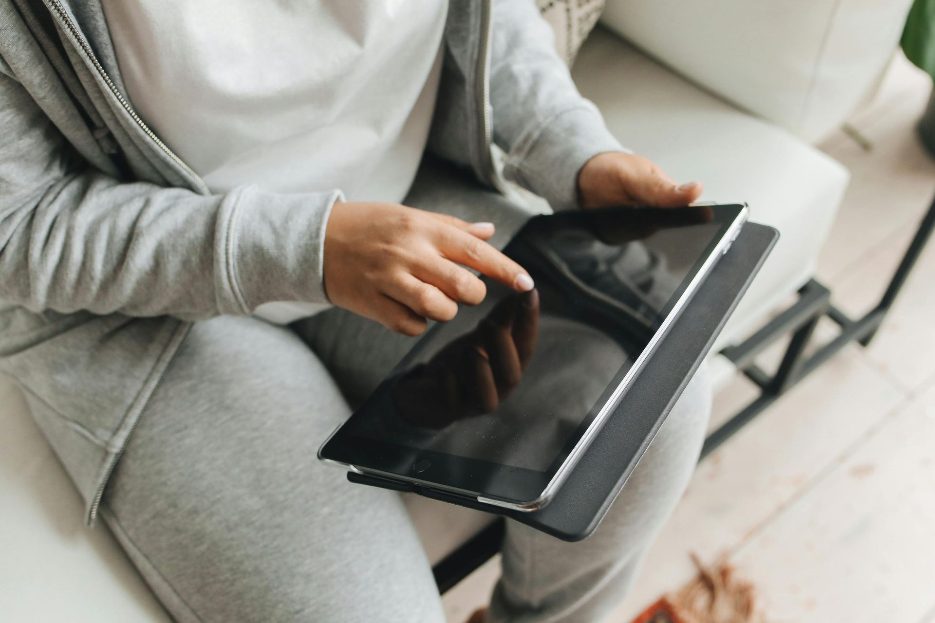 Person in gray sweatsuit using a tablet, sitting on a white couch.