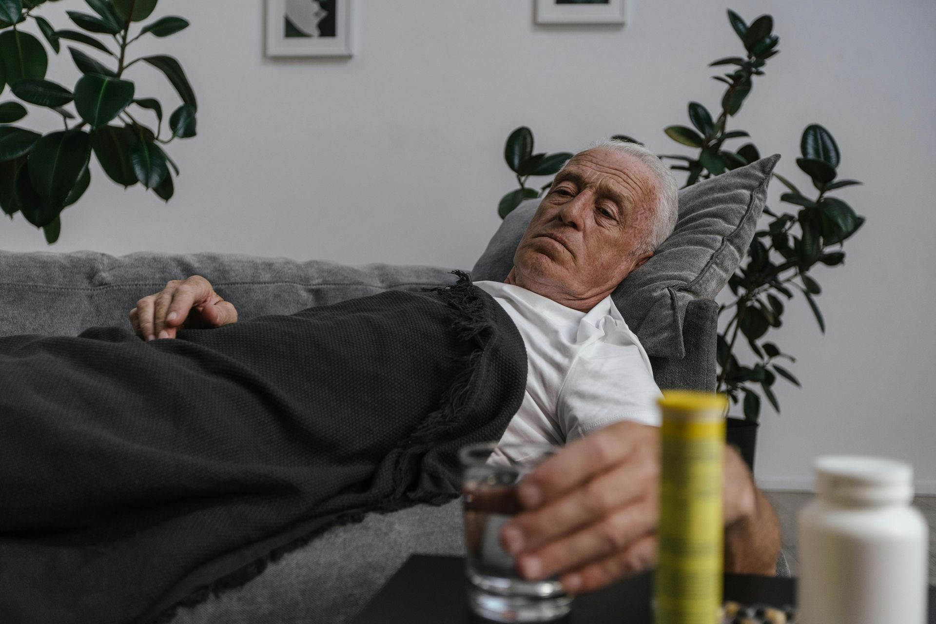 Elderly man lying on a couch, appearing ill. He has a glass of water and medication within reach.