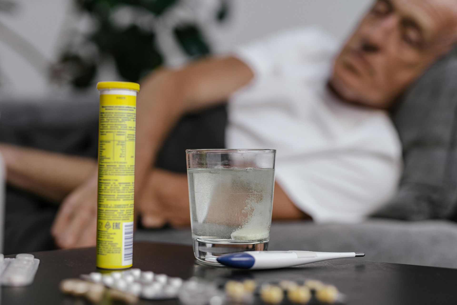 A sick person rests on a couch next to medicine, a glass of water, and a thermometer on a table.
