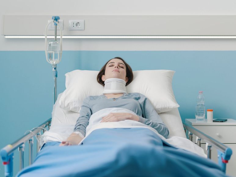 Woman in hospital bed wearing a neck brace; an IV drip is next to her, and a table with medication and a water bottle is nearby.
