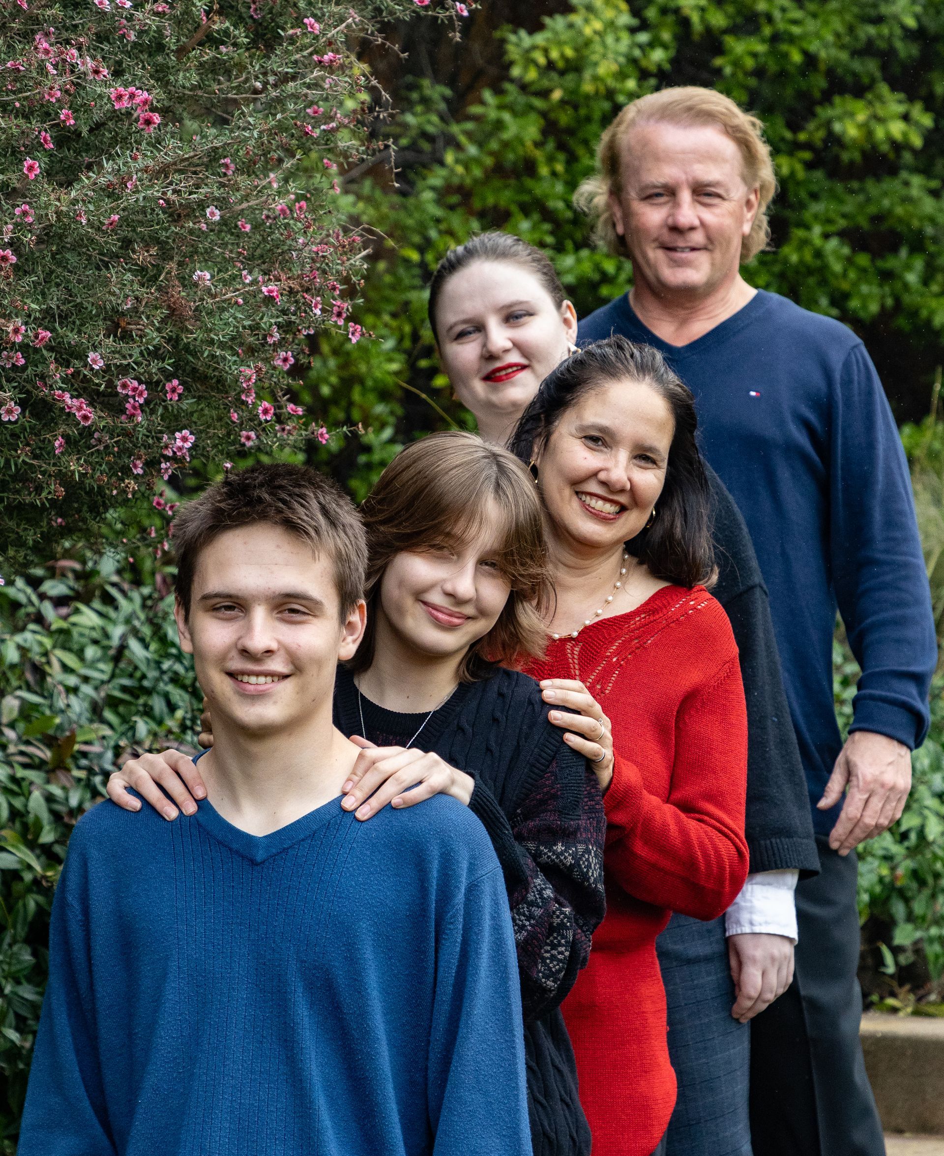 Family of five standing in front of greenery; smiles, a red sweater, blue sweaters, and a casual setting.