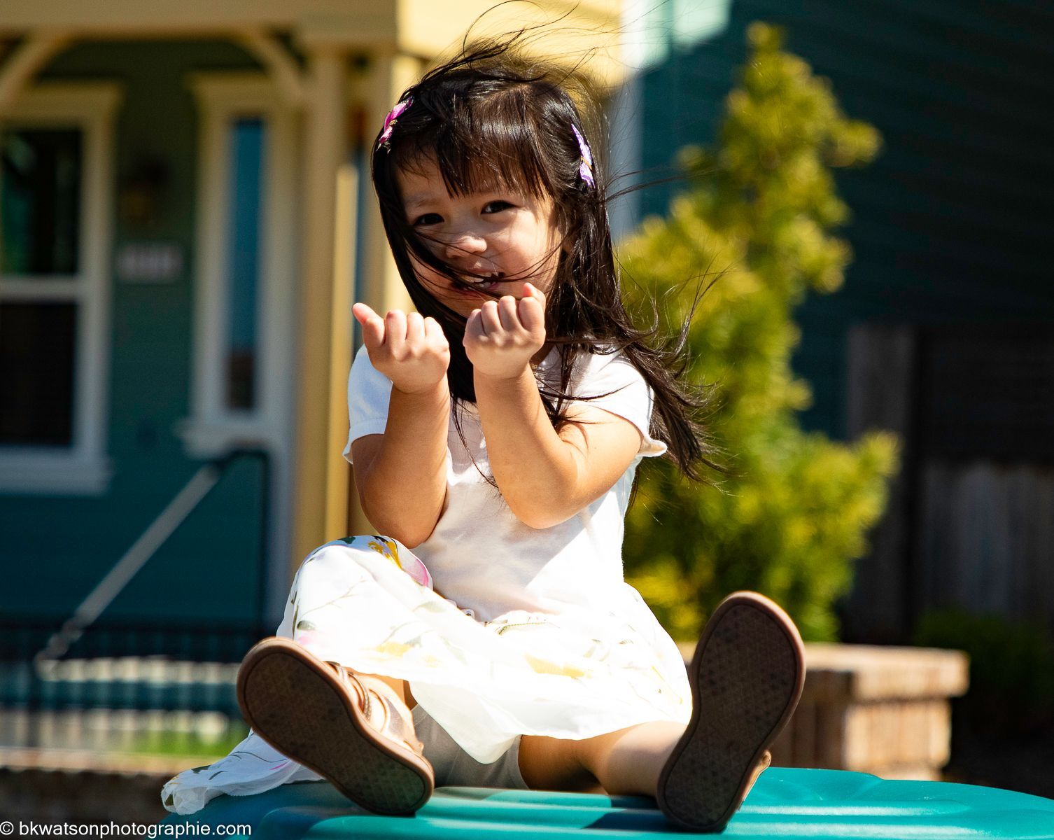 Young child sitting outdoors, smiling, with hands up, brown shoes, light clothing.