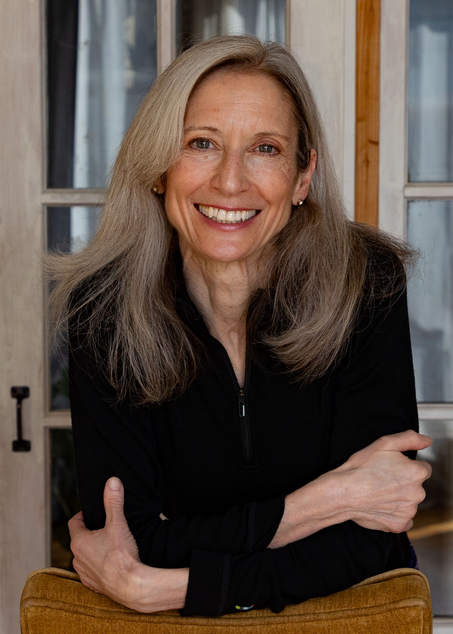 Woman with long gray hair smiles, leaning on an upholstered chair, wearing a black jacket.