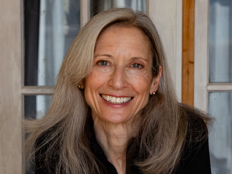 Woman with long gray hair smiling, in front of a window. She wears a black shirt.