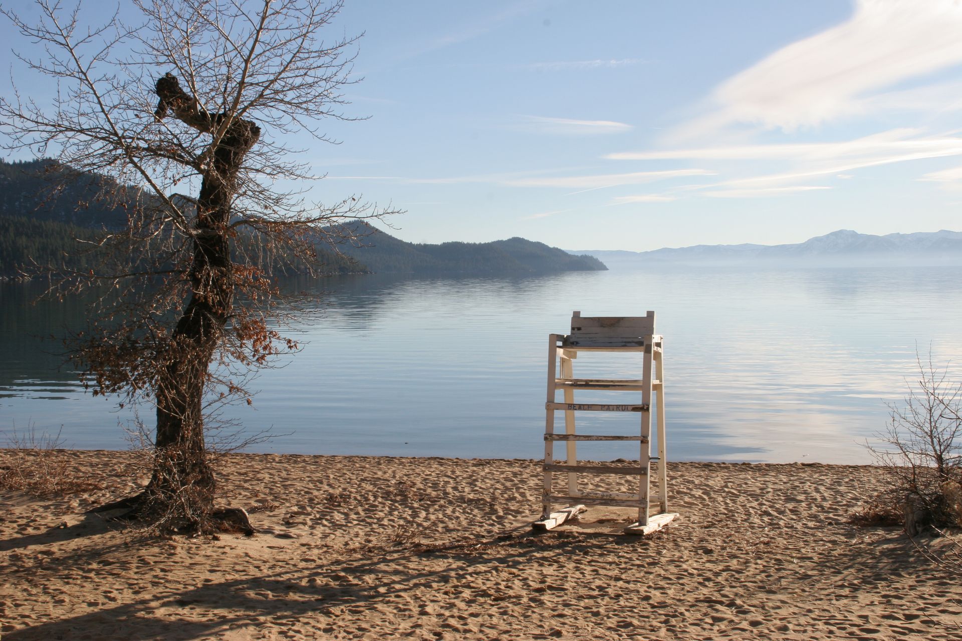 Sandy beach with a lifeguard stand, bare tree, and calm lake under a clear sky. Mountains in the distance.