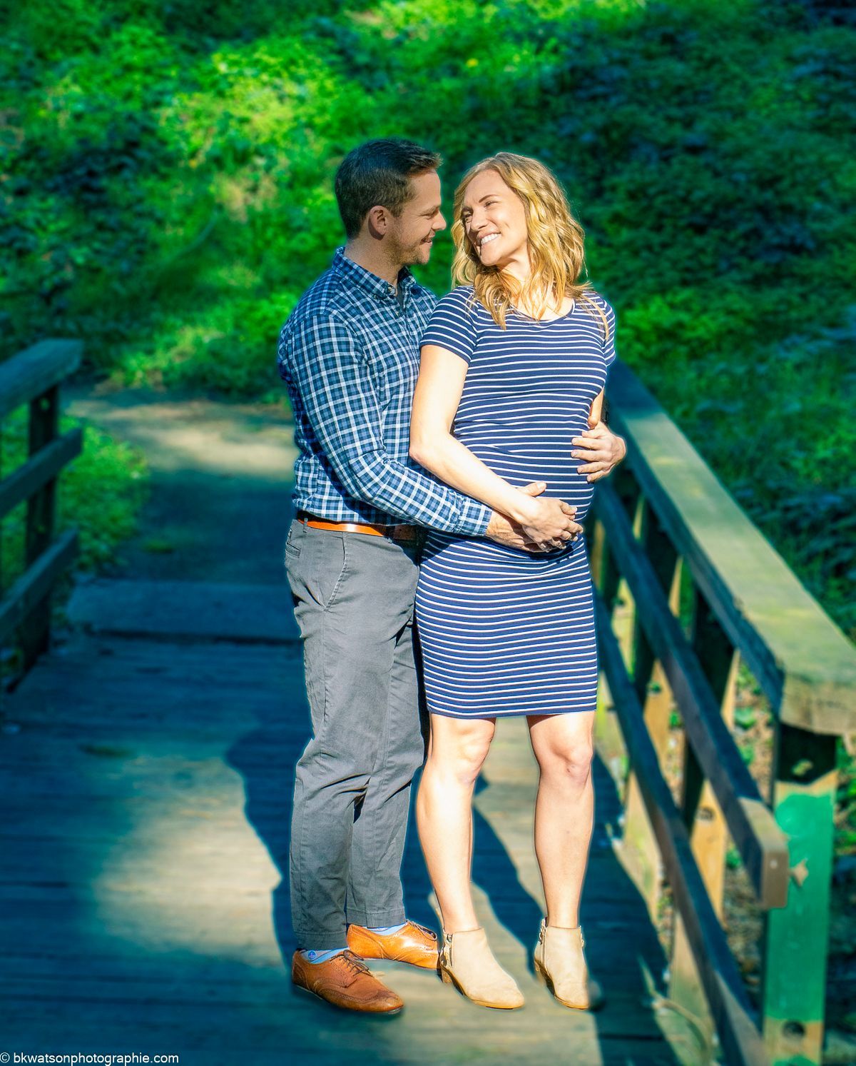 Couple embraces on a wooden bridge in a green forest; woman in blue striped dress, man in plaid shirt.