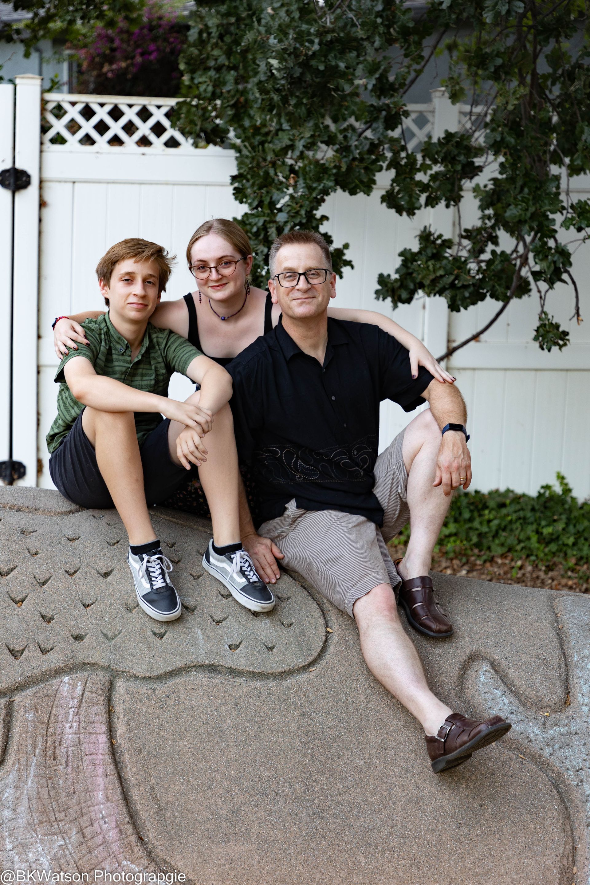 A family of three sitting on a rock wall, smiling. A boy in green and a girl with glasses embrace a man in black and shorts.