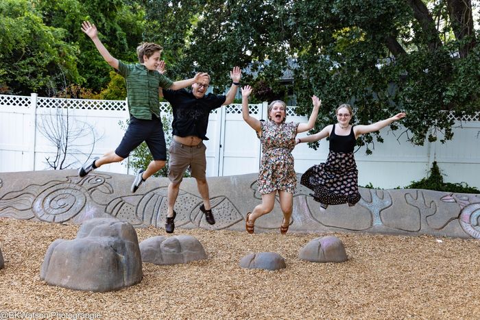 Four people jumping joyfully in a park; rocks and wood chips on the ground, trees and fence in the background.