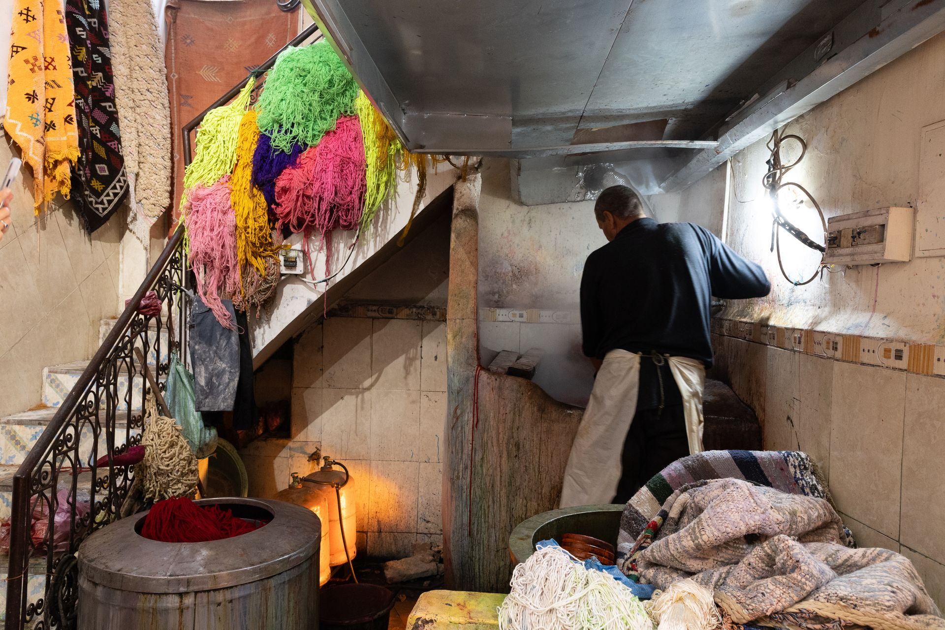 Man dyeing wool in a workshop, colorful skeins hanging near staircase.