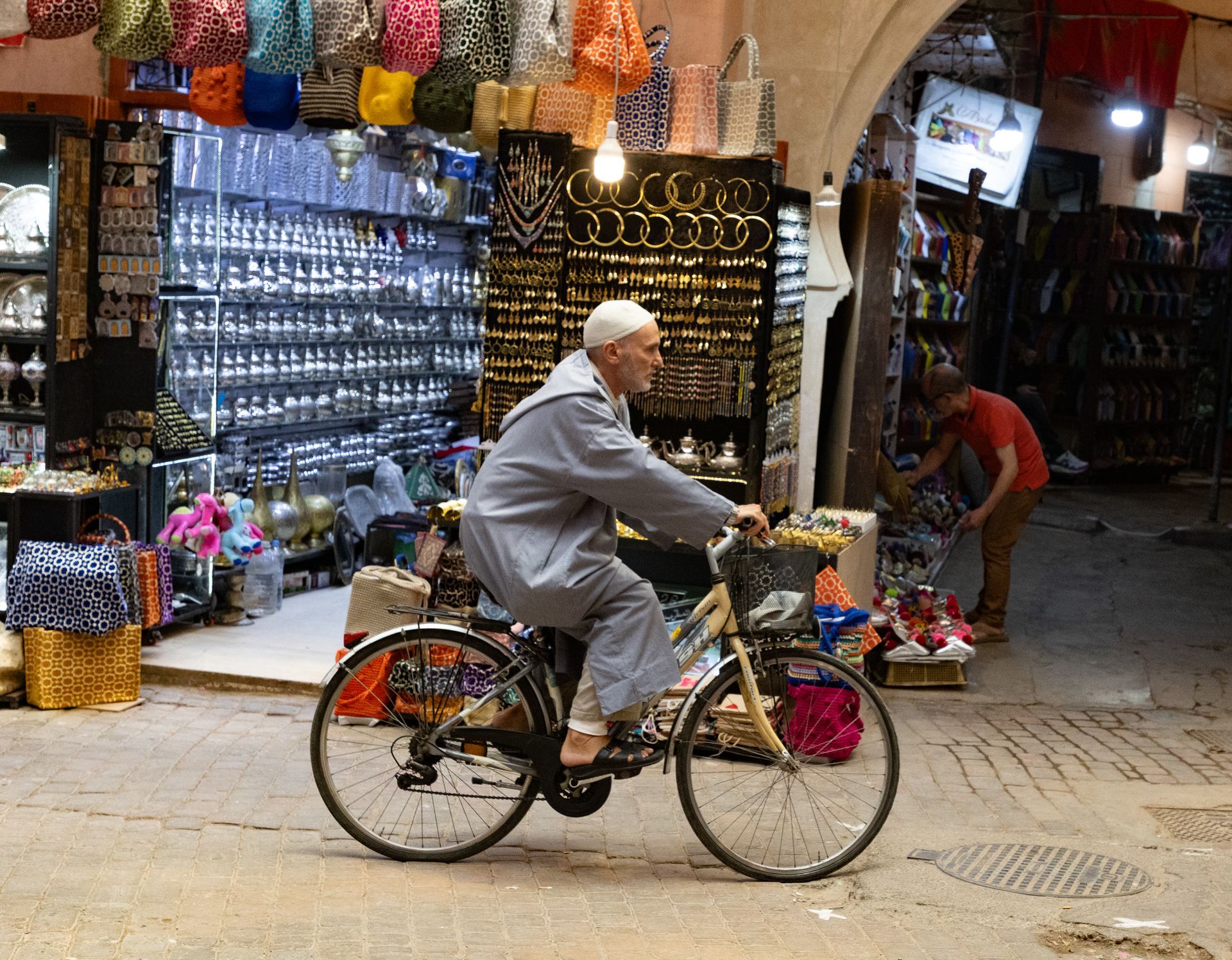 An elderly man on a bicycle rides through a bustling marketplace, colorful goods on display.