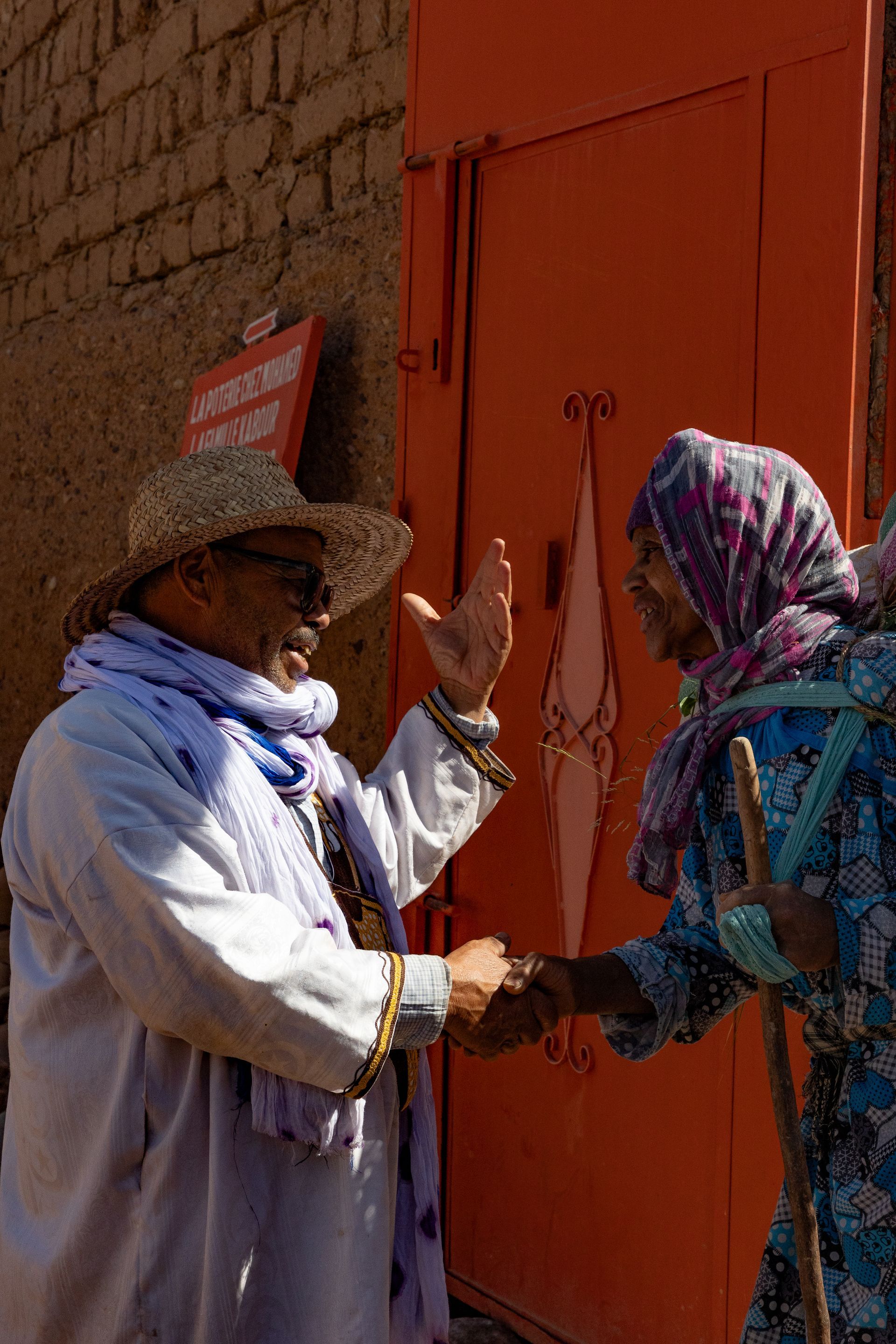 Two people greet, one wearing a straw hat and scarf, the other a headscarf, by an orange door.