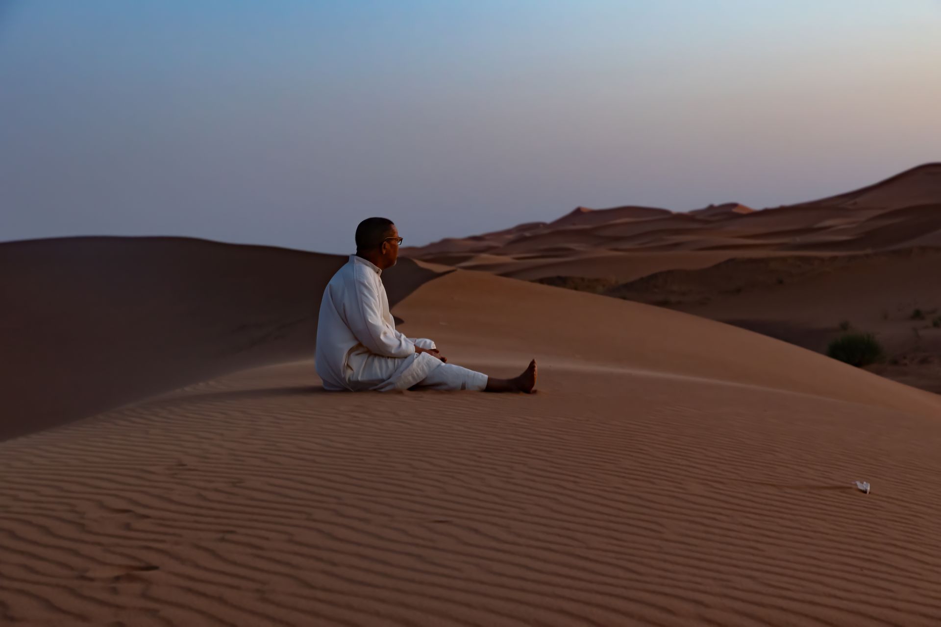 Man seated on a desert dune at dusk, wearing white robes, gazing at the horizon.