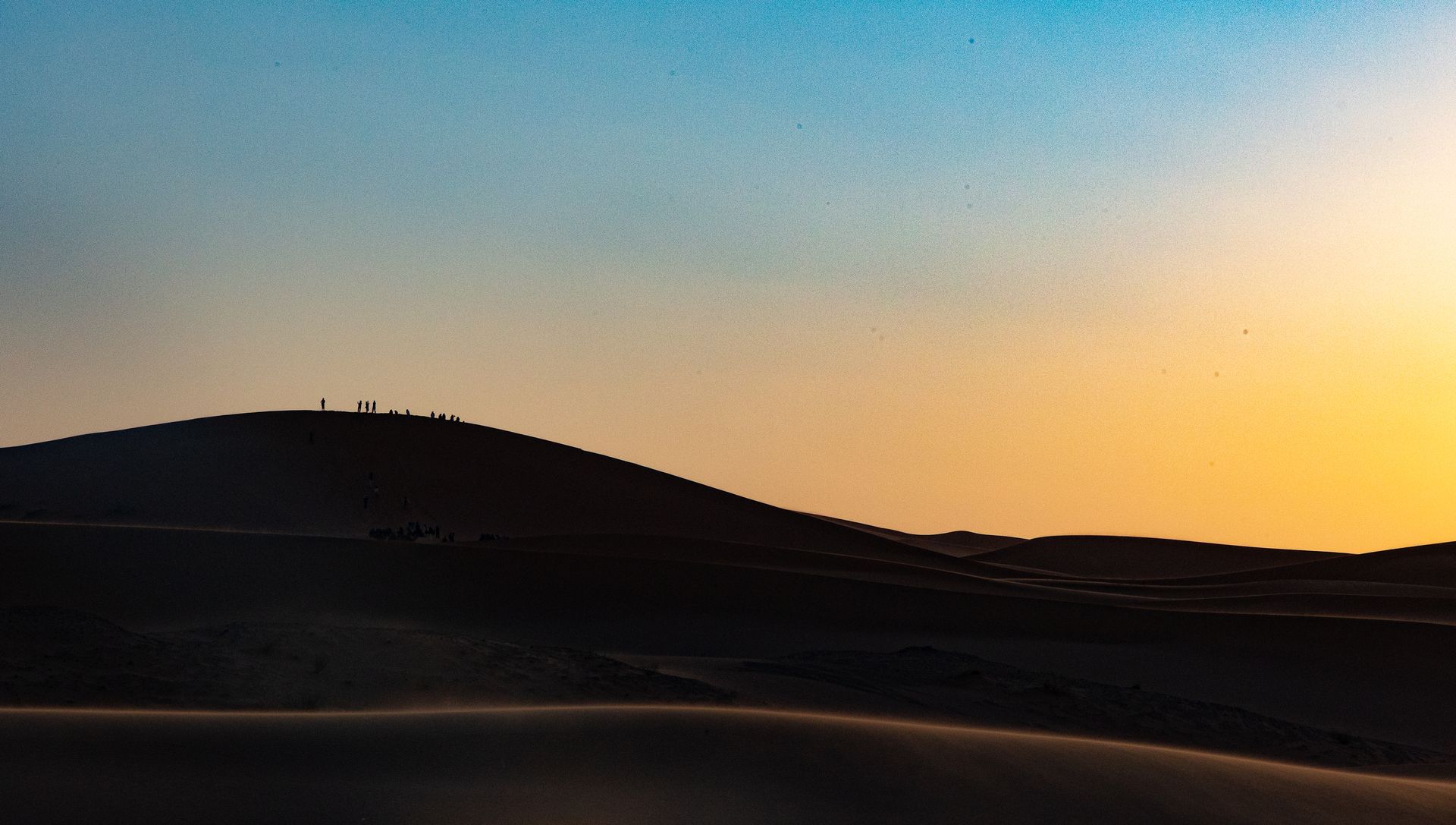 Desert dunes silhouetted against a colorful sunset sky; people on a distant ridge.