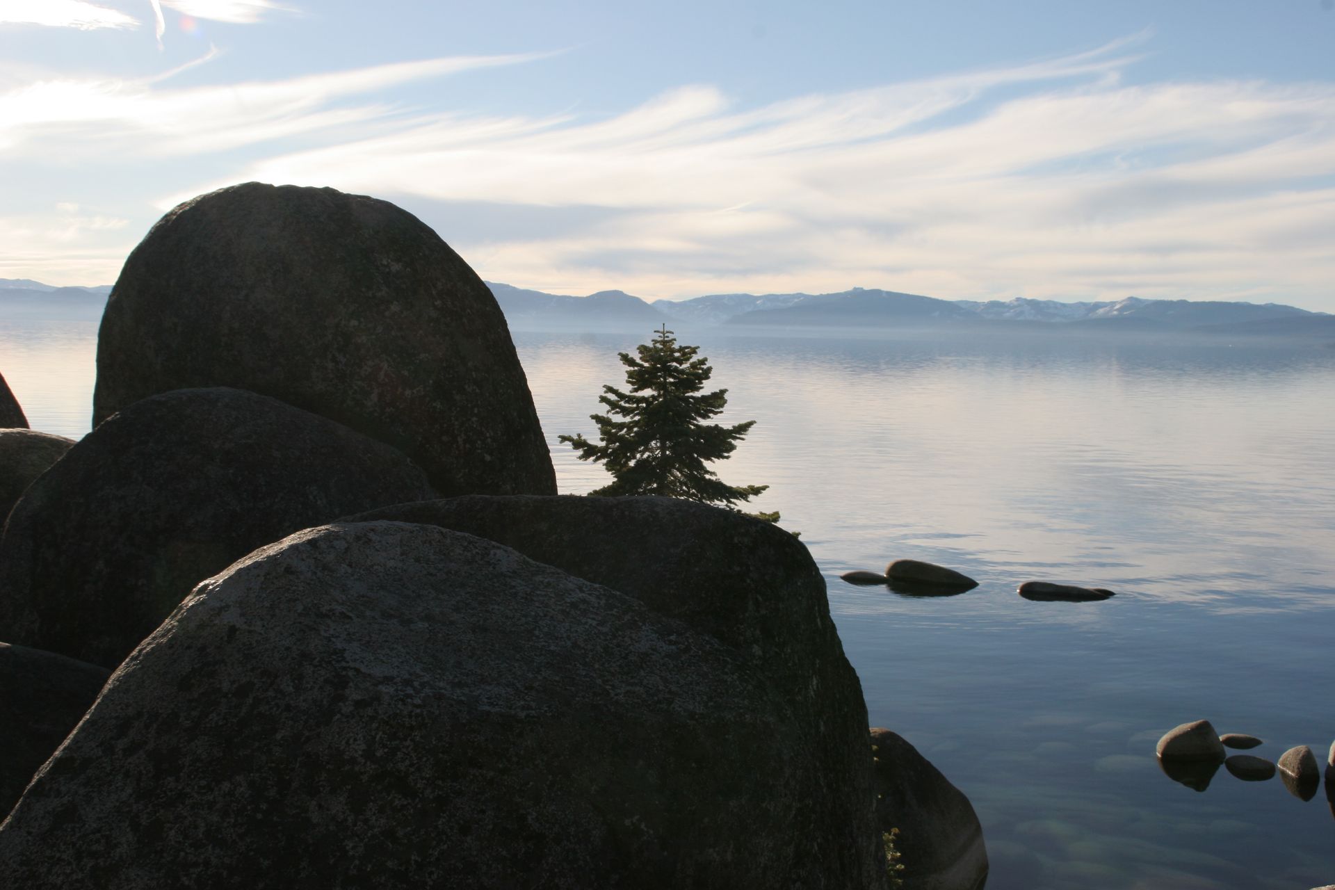 Large dark rocks in foreground, with calm lake and distant mountains under a cloudy sky.