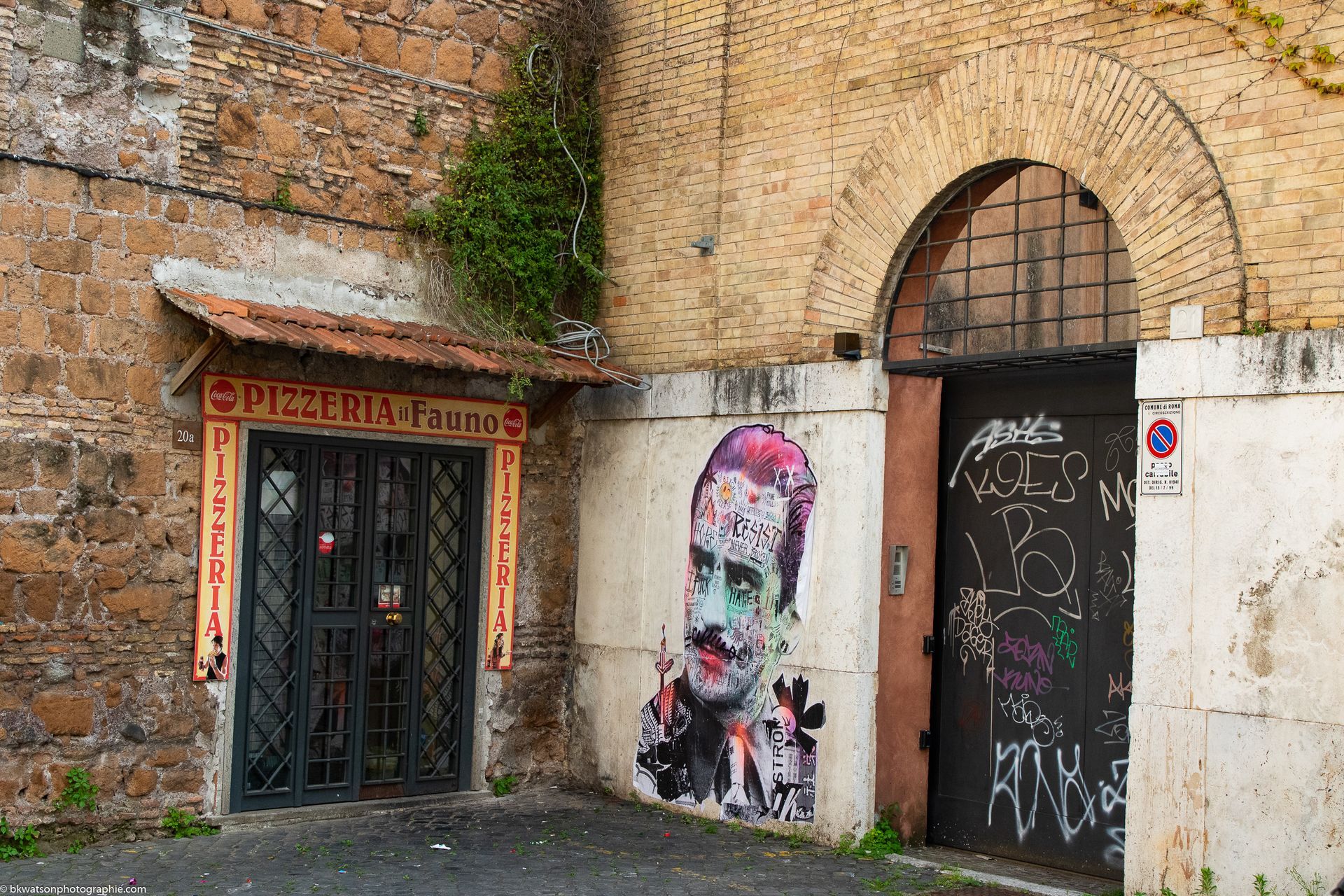 Exterior of buildings with a doorway, graffiti portrait, and arched entrance. Dark, worn facade with greenery.
