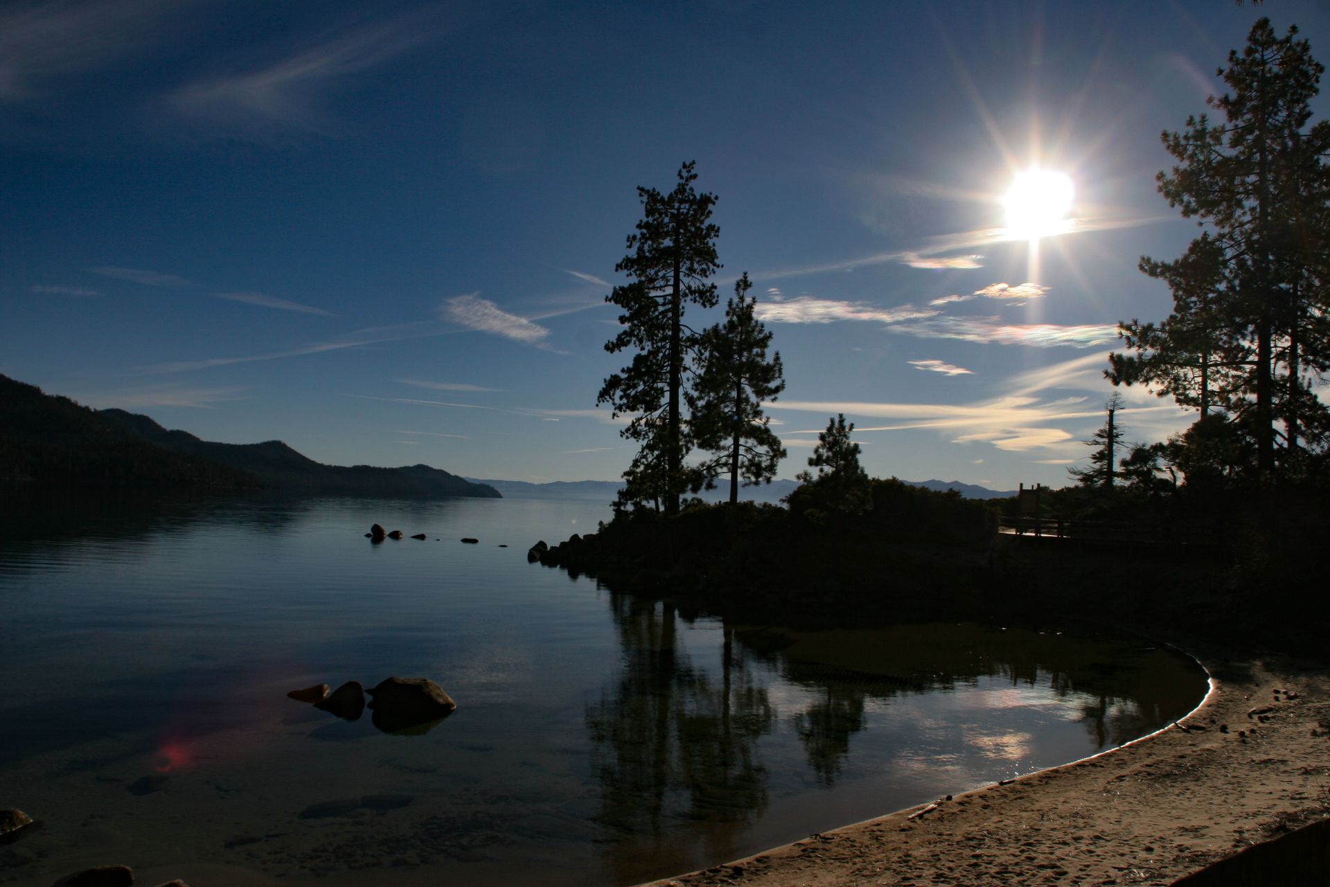 Sun over calm lake, silhouetted trees on shore reflect in water.