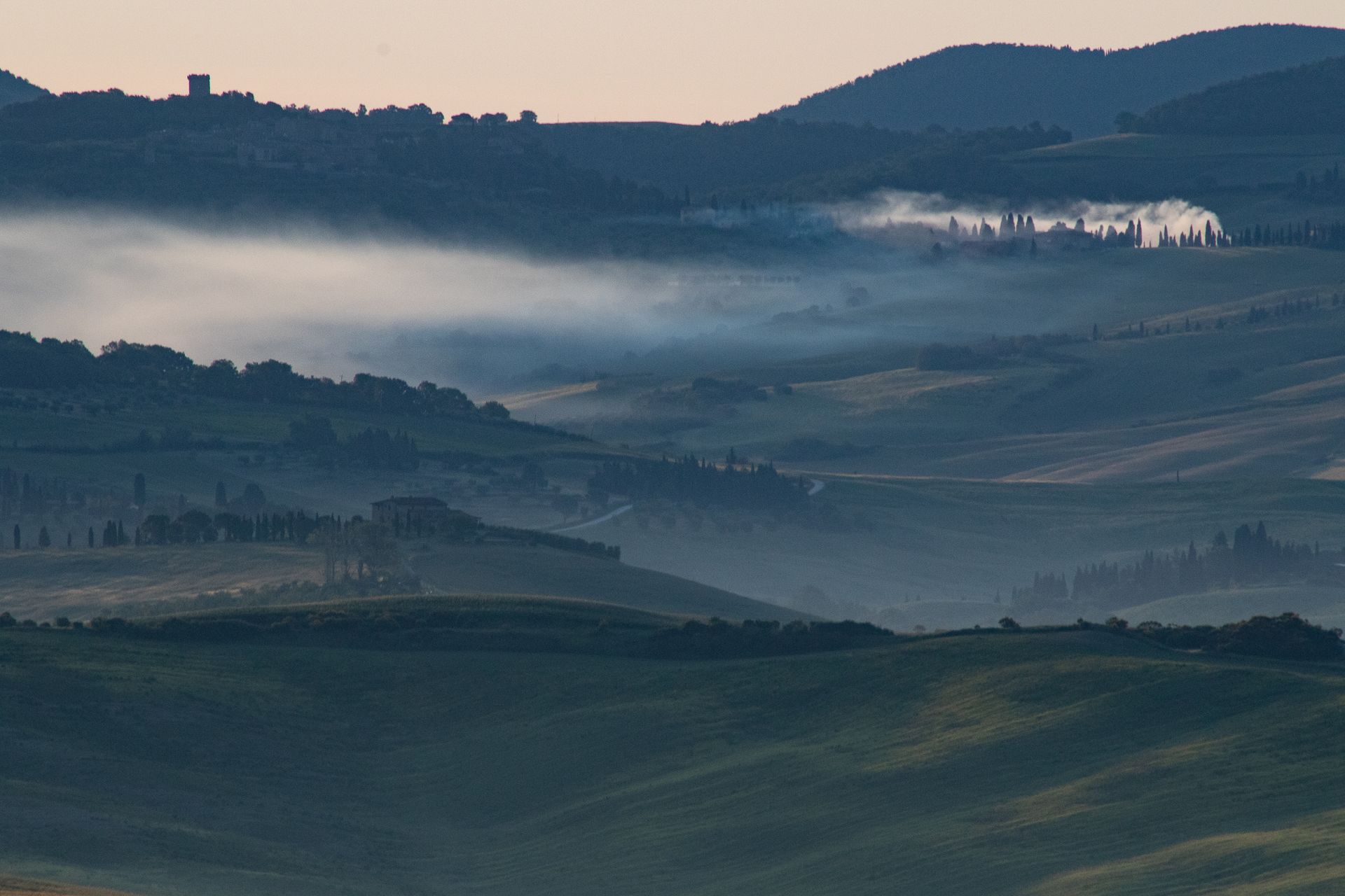 Rolling green hills in a misty valley with distant buildings, silhouetted under a pale sky.