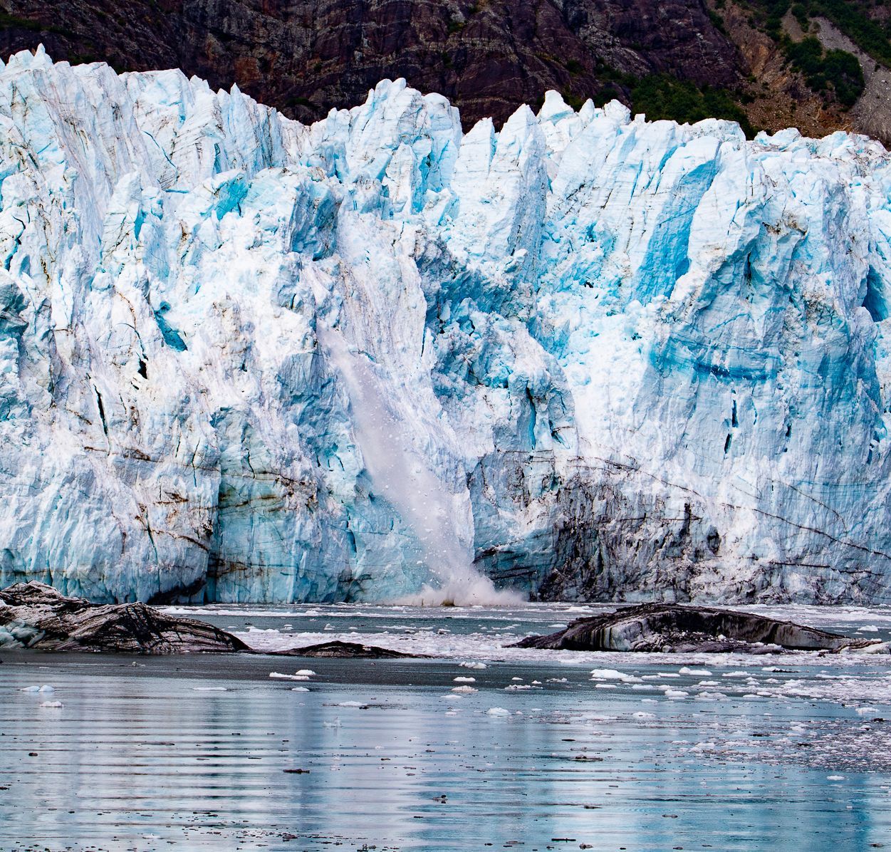Glacier calving into the water; icy blue and white ice with a dark rock backdrop.