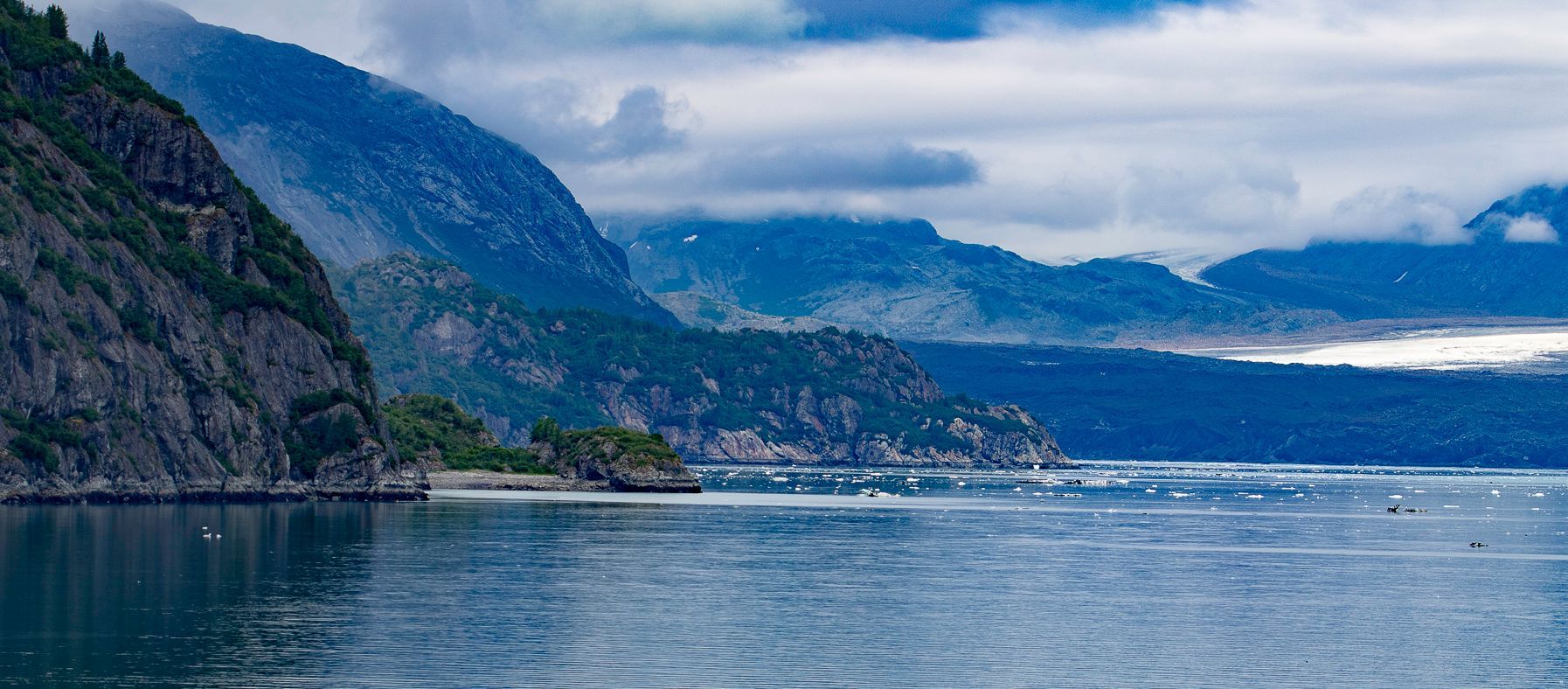 Mountains meet the ocean under a cloudy sky. The landscape is mostly blues and greens.
