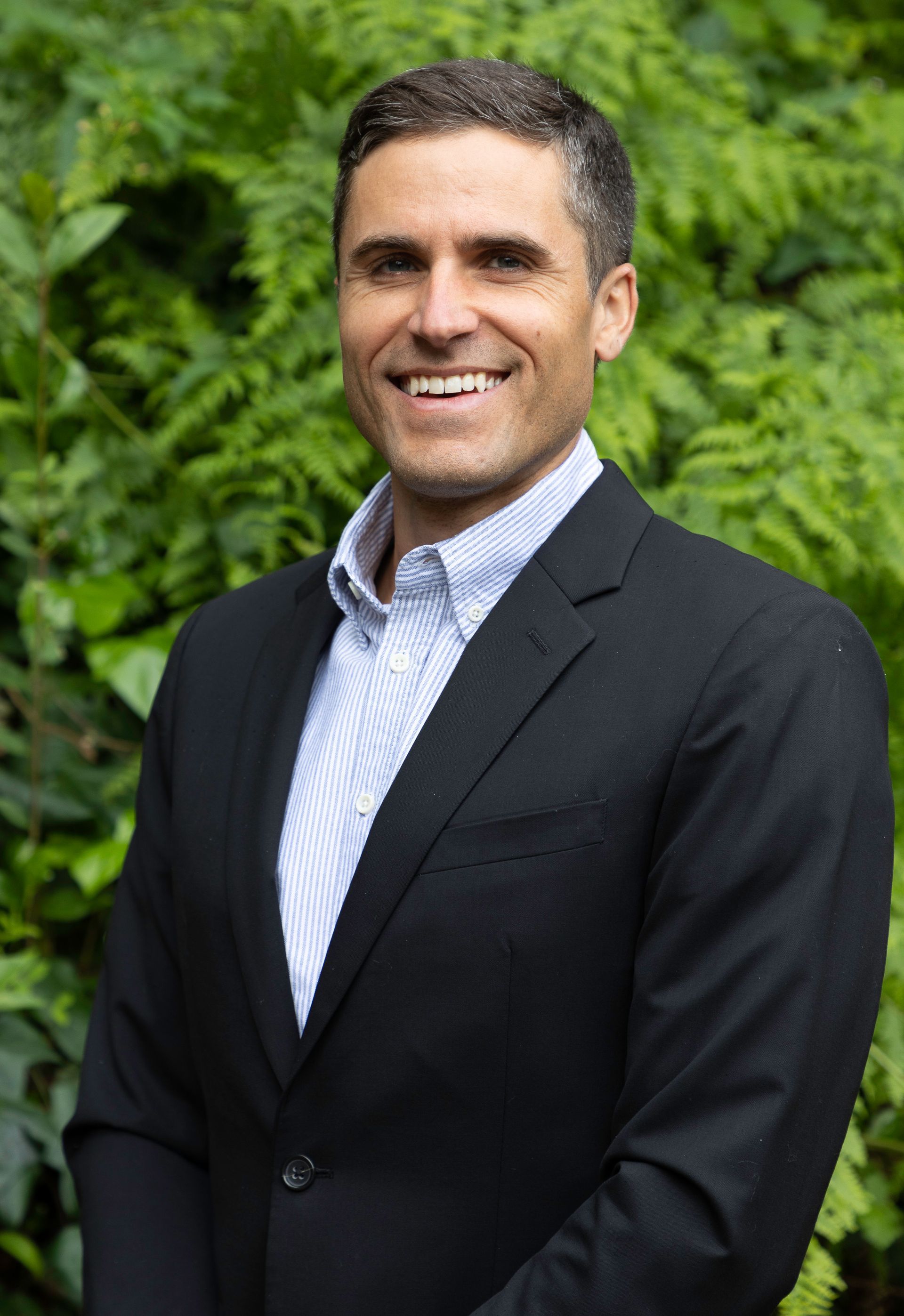 Man in a black blazer and blue shirt smiling in front of green foliage.