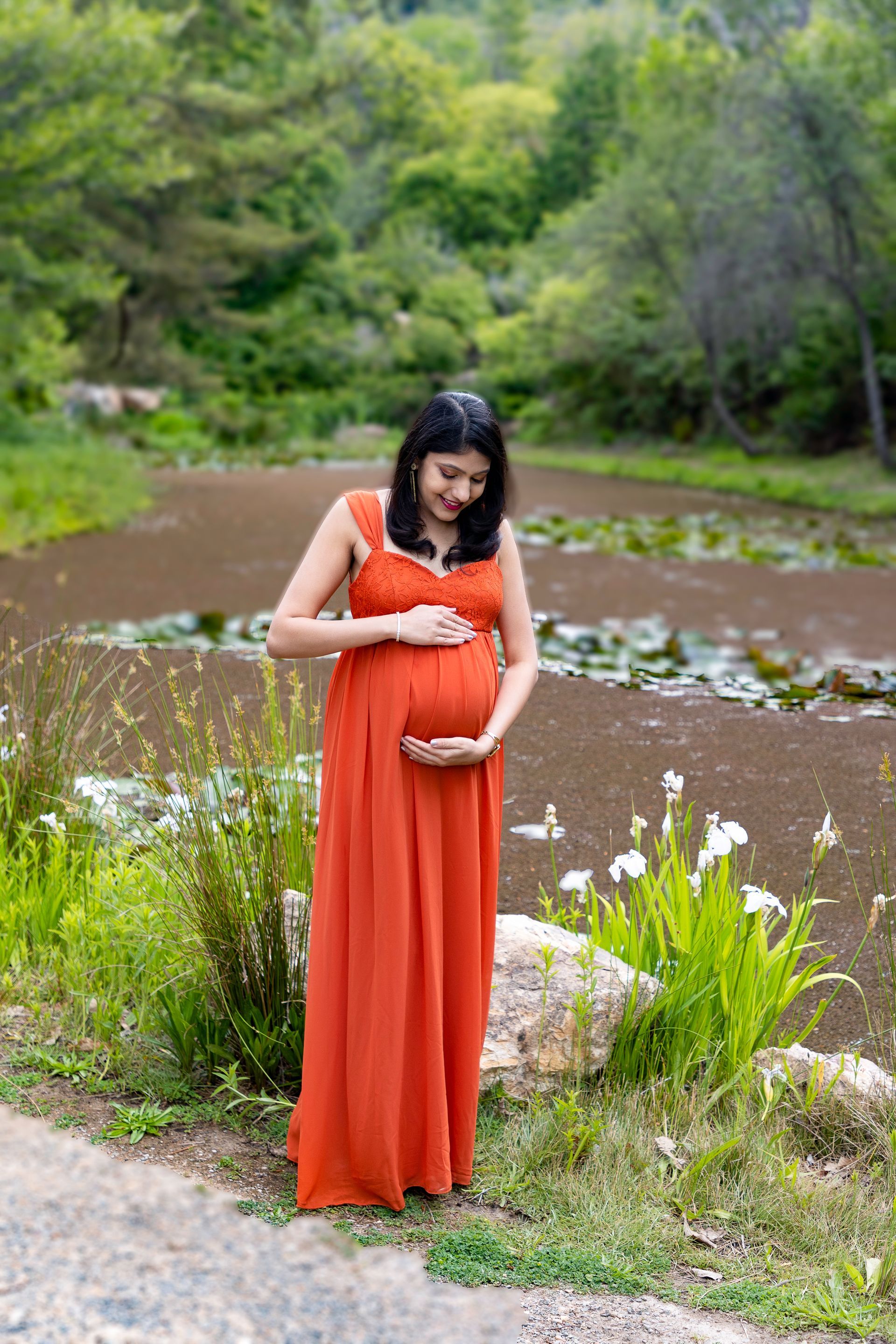 Woman in orange dress cradling pregnant belly, standing near water with green foliage.