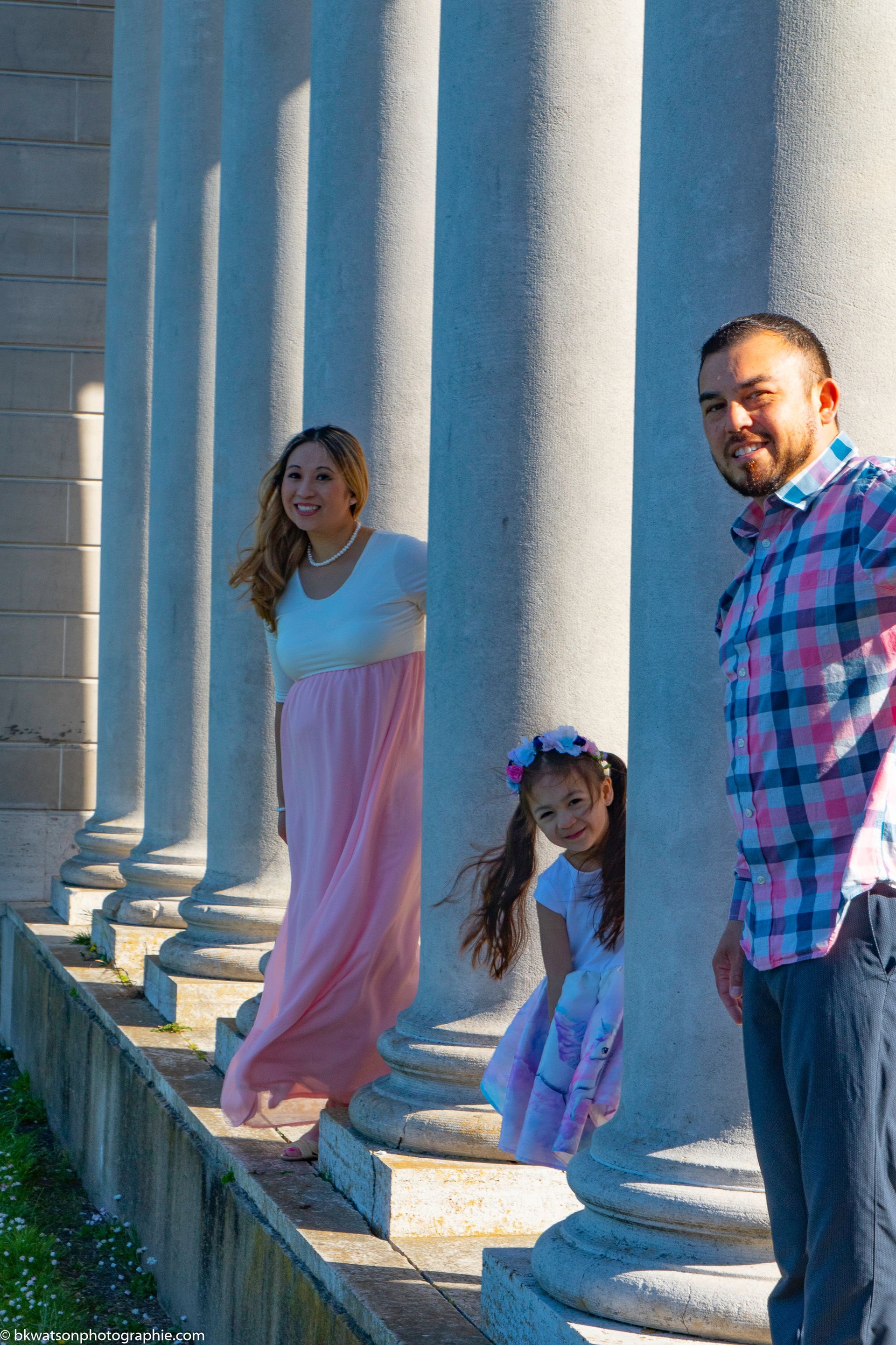 Family of three posing near tall white columns. Woman in pink dress, child in dress with a flower crown, man in plaid shirt.