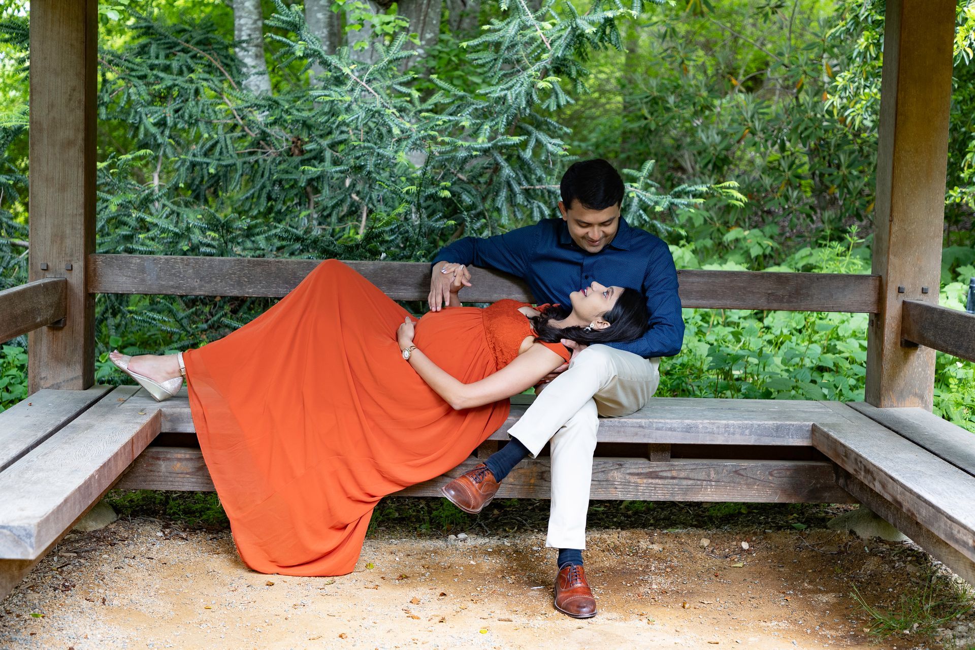 Couple in gazebo, pregnant woman in orange dress, man in blue shirt, resting, smiling.