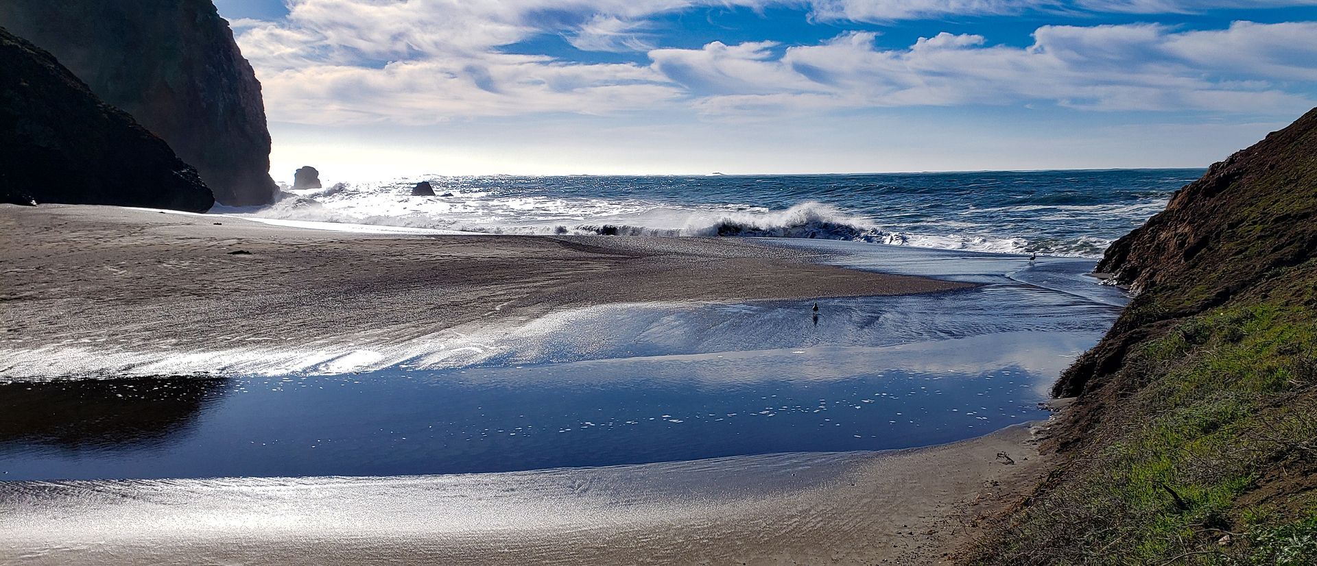 Ocean waves crash onto a beach, with a pool of water reflecting the sky. Cliffs frame the view.