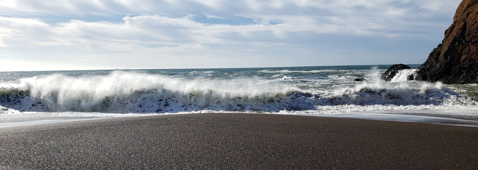 Waves crashing on a dark, pebbly beach under a cloudy sky.
