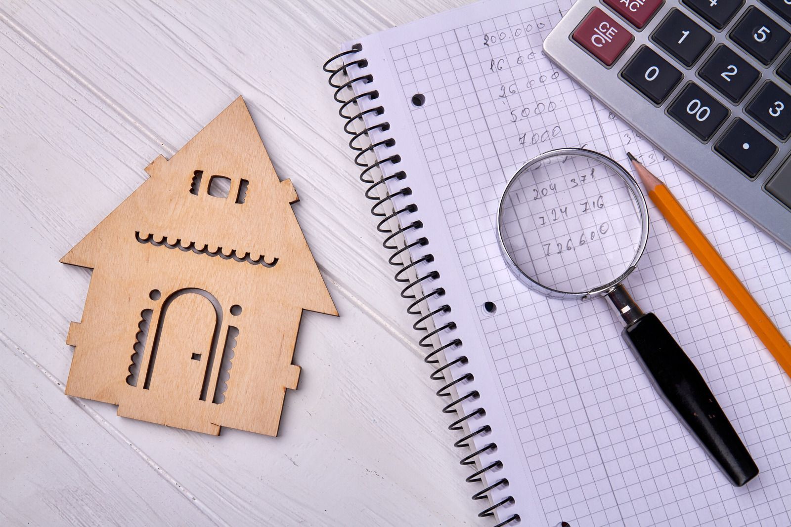 A wooden house is sitting on top of a notebook next to a magnifying glass and a calculator.