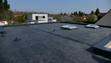 Man on a gray shingle roof, surrounded by green trees.