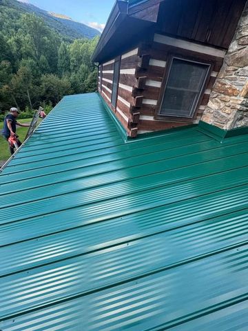 Green metal roof on a log cabin with workers, trees, and mountains in the background.
